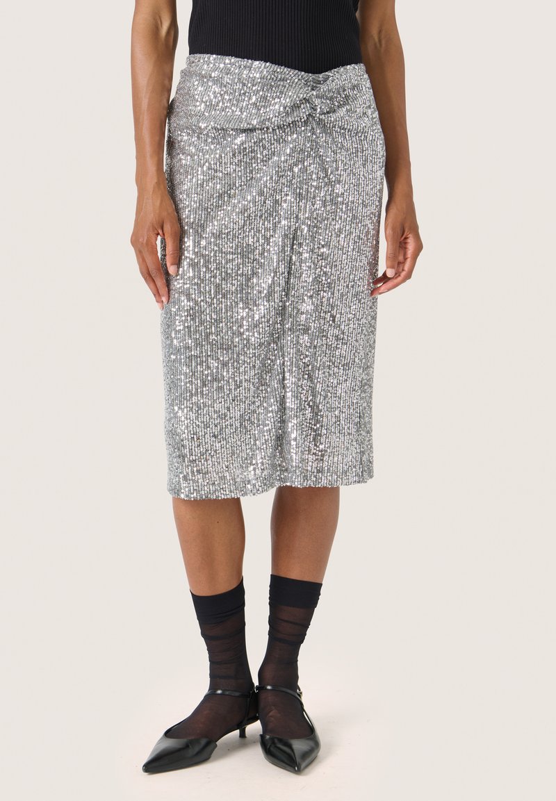 Zilveren paillettenrok met een geknoopte taille, met verticale ribbelstructuur. Draagt het met zwarte doorschijnende sokken en puntige zwarte schoenen.