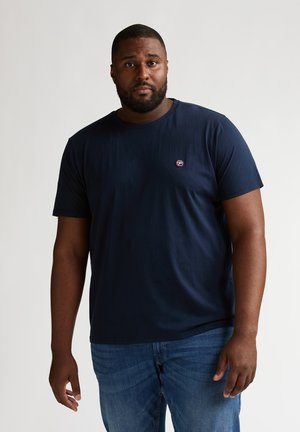 Tricou din bumbac albastru marin, cu mâneci scurte, decolteu rotund și un mic logo pe pieptul stâng. Purtat cu blugi albaștri.