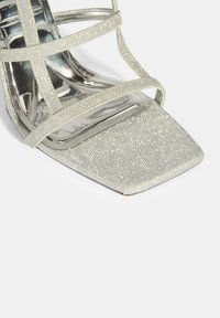 Silberne Glitzer-High-Heel-Sandalen mit dünnen Riemen und einem quadratischen Zehendesign. Das Material hat eine strukturierte, funkelnde Oberfläche.