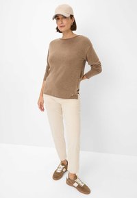 Brauner Strickpullover mit langen Ärmeln, cremefarbene Cordhosen und braune Turnschuhe. Die Mütze ist beige mit einem hellblauen Akzent.