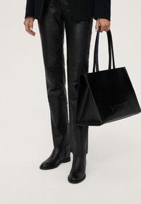 Sac à main en cuir noir avec logo embossé, assorti à un pantalon en cuir noir avec des motifs texturés, et des bottines en noir assorti.
