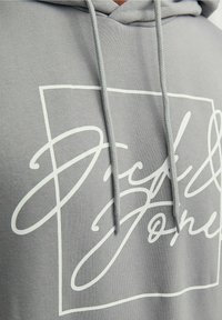Sudadera gris hecha de una tela suave, con un gran diseño de logo blanco dentro de un contorno rectangular. Los cordones son visibles en el cuello.