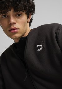 Giacca in pile nera con zip, con un logo Puma bianco sul lato sinistro, e un maglione nero a coste sotto.