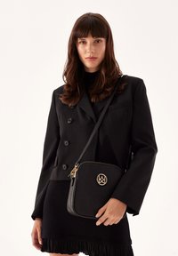 Borsa a tracolla in pelle nera con forma quadrata, dettaglio del logo oro, chiusura zip e tracolla regolabile. La superficie ha una finitura texturizzata.