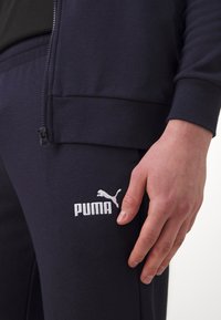 Marinblå atletisk träningsdräkt med en jacka med dragkedja och sweatpants. Vit broderad logotyp på byxorna, mjuk textur och fin passform.