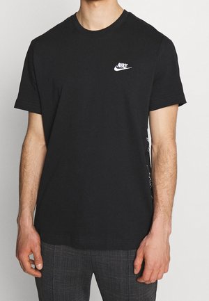 Mężczyzna ubrany w czarną koszulkę Nike z białym logo na piersi i szare spodnie w kratę, stojący na tle jednolitego białego tła.