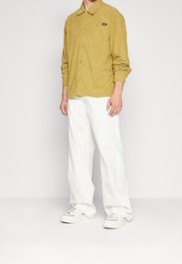 Camicia di velluto a coste color senape con colletto e chiusura a bottoni, abbinata a pantaloni bianchi larghi e sneakers bianche. Dettaglio del logo nero visibile.
