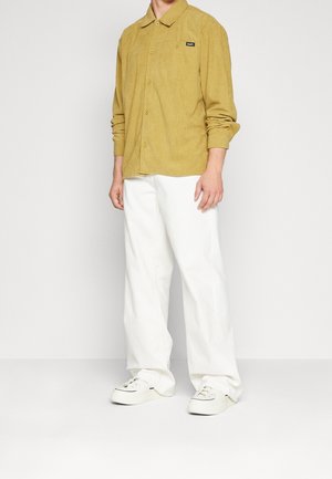 Camicia di velluto a coste color senape con colletto e chiusura a bottoni, abbinata a pantaloni bianchi larghi e sneakers bianche. Dettaglio del logo nero visibile.