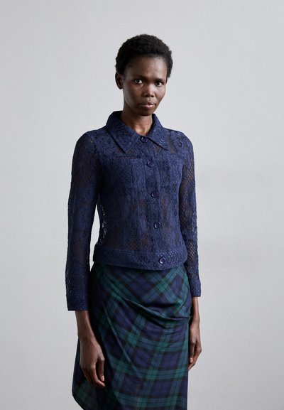 Marineblaue Spitzenjacke mit langen Ärmeln, einem Kragen und lila Knöpfen. Das Model trägt einen karierten Rock in dunklen Grün- und Marineblau-Tönen.