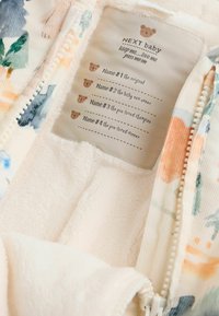 Prenda para bebé con un estampado floral y un forro de forro polar color crema. Cuenta con una cremallera y una etiqueta de cuidado con espacios para nombres y un gráfico de un osito de peluche.