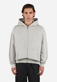 Sudadera con cremallera en un gris claro, hecha de material suave, que cuenta con un bolsillo frontal y un logo en el lado izquierdo del pecho, con un corte relajado y capucha ajustable.
