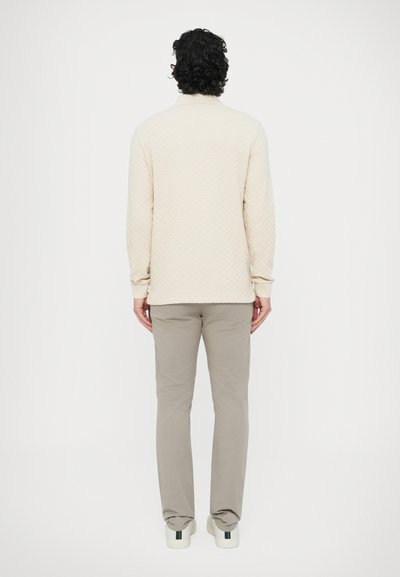 Ανοιχτό μπεζ πουκάμισο μακρυμάνικο με καπιτονέ σχέδιο και γιακά, συνδυασμένο με ανοιχτό γκρι παντελόνι και άσπρα αθλητικά παπούτσια με έγχρωμη ρίγα στη φτέρνα.