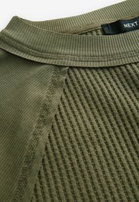 Tissu texturé vert olive avec un design cannelé, encolure lisse et piqûres visibles. Étiquette affichée à l'intérieur du col.