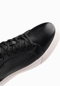Baskets en cuir noir avec lacets plats, bout arrondi et semelle en caoutchouc blanc. Présentent des coutures et des détails perforés sur le côté.