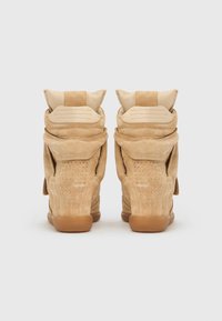 Paire de baskets compensées en daim beige avec des clous décoratifs sur les talons, des cols rembourrés repliables et des semelles en caoutchouc sur fond blanc.