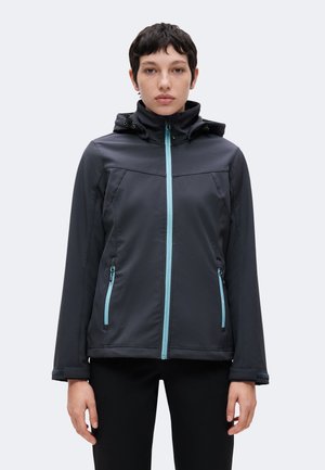 Jeune femme portant une veste noire à capuche avec des fermetures éclair bleu clair, debout face à un fond clair uni.