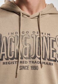 Beige Baumwoll-Hoodie mit großem schwarzem Grafikdruck, der "Jack & Jones" zeigt, mit "INDIGO DENIM" und "SINCE 1990" darunter.