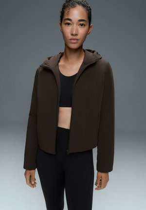 Frau mit lockigem Haar trägt eine dunkle kurze Jacke über einem schwarzen Sport-BH und Leggings vor einem einfarbigen grauen Hintergrund.
