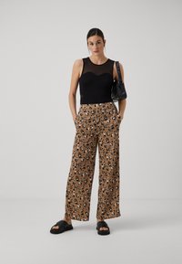 Svart ermeløs topp med mesh-overlegg, brune vide bukser med svart og hvit leopardmønster, parret med svarte sandaler og en håndveske.