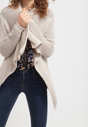 Femme portant un cardigan beige en tricot par-dessus un chemisier à fleurs et un jean bleu foncé, tenant le cardigan fermé avec les deux mains.