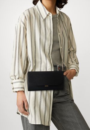 Persona che indossa una camicia a righe beige con bottoni sopra una maglietta grigia e jeans, con una borsa a tracolla nera con catena dorata.