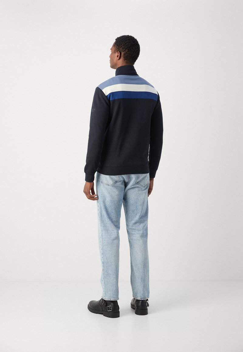 Pull bleu marine avec des rayures horizontales en bleu clair et bleu foncé au dos ; porté avec un jean bleu clair et des bottes noires.