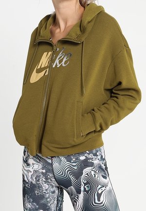 Donna che indossa una felpa con zip Nike verde oliva e leggins con motivo astratto in bianco, nero e grigio, con le mani nelle tasche della felpa.