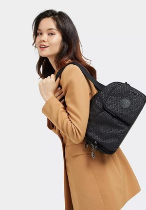 Black patterned backpack with a textured finish, two front pockets, and adjustable strap, worn over a tan coat.