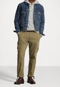 Giubbotto di jeans, felpa grigia con cappuccio e pantaloni cargo oliva. Stivali marroni con lacci. Caratteristiche includono dettagli con tasche sui pantaloni e texture a contrasto.