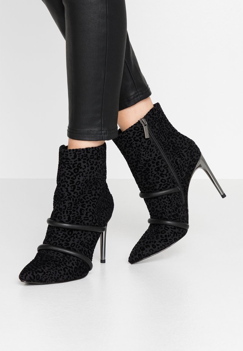 Bottines noires à imprimé léopard, talons hauts, fermeture éclair sur le côté et brides décoratives, portées avec un pantalon noir ajusté.