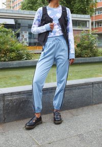 Ljusblå sweatpants med svarta sidostråk, kombinerat med en svart väst över en mönstrad långärmad skjorta och svarta skor.