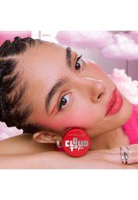 Junge Frau mit geflochtenem Haar legt ihren Kopf auf den Arm, trägt rosa Rouge und roten Eyeliner, hält einen roten Make-up-Behälter von Maybelline Cloud Topia.