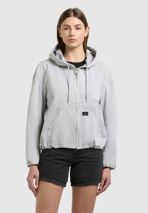 Jeune femme portant une veste à capuche zippée gris clair et un short en denim noir, debout devant un fond gris uni.
