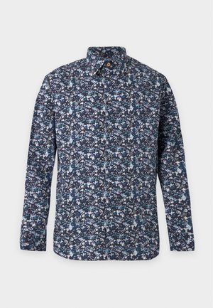 En ung mand står med den ene hånd i lommen iført en blå blomsterprintet langærmet skjorte, mørke denimshorts, brune sko og grå sokker.