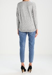 Harmaa pitkähihainen paita, jossa on teksturoitu kangas ja koristeelliset olkapainikkeet, yhdistettynä vaaleansinisiin kapeisiin farkkuihin ja mustiin korkokenkiin.