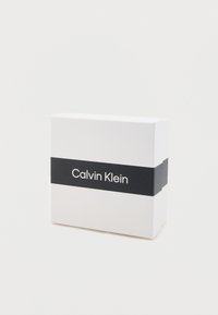 Baltas stačiakampis dėžutė su juoda horizontalia juosta, kurioje balta spalva užrašyta "Calvin Klein". Lygus paviršius, minimalistinis dizainas.