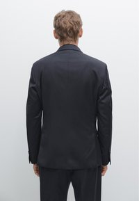 Dunkler Nadelstreifenanzug mit Reverskragen, taillierter Passform und einfachem Rückenschlitz, von hinten vor einem schlichten hellen Hintergrund gezeigt.