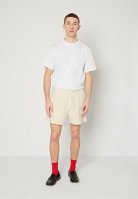 T-shirt blanche à manches courtes, shorts beiges avec une texture douce et une ceinture élastique, chaussettes rouges et chaussures noires à enfiler.