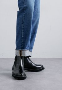 Black Chelsea boots with a glossy finish, rubber sole, paired with cuffed blue denim jeans showing contrasting stitching.