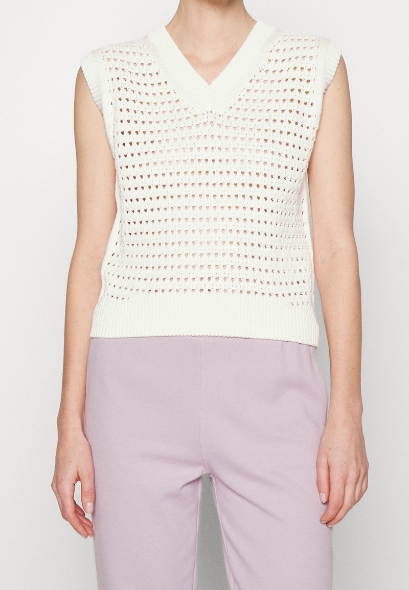 Gilet blanc sans manches en tricot avec un motif ajouré sur les bras nus, associé à un pantalon taille haute violet clair porté par une personne.