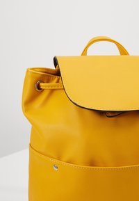 Mochila de cuero amarillo con forma redondeada, cierre de solapa, parte superior con cordón y un bolsillo frontal con detalle metálico a presión.