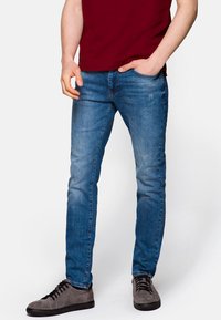 Světle modré denimové džíny s úzkým střihem, s jemnými odřenými detaily. Nosené s šedými botami a vínovou košilí.