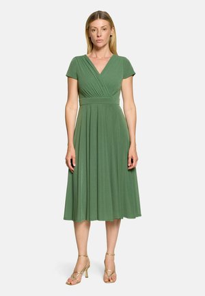 Femme debout portant une robe midi verte à manches courtes avec un col en V et des sandales dorées à talons hauts à brides sur un fond uni.
