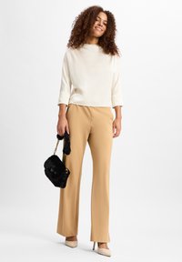 Cremetop mit 3/4-Ärmeln, beigen Hosen und schwarzer, pelziger Handtasche. Das Model trägt beigen Absätze; das Outfit hat ein klares, minimalistisches Design.
