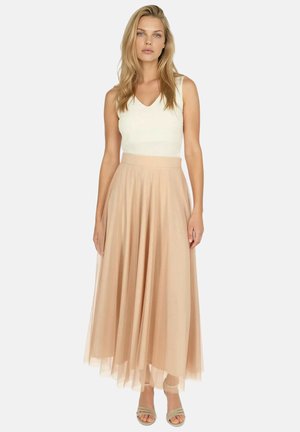 Kobieta ubrana w biały, bezrękawnik i długą, zwiewną beżową spódnicę z beżowymi butami na obcasie z odkrytymi palcami, stojąca na tle jednolitego tła.