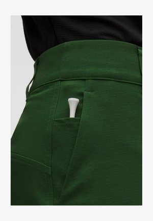 Približan posnetek zelene žepne hlačnice z delno vidnim belim golfskim klinčkom znotraj, nošen s črnim zgornjim delom.