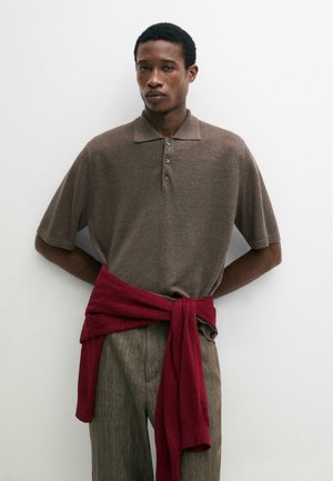 Homme portant un polo marron à manches courtes avec un pull rouge noué autour de la taille et un pantalon marron à fines rayures, debout contre un mur blanc.
