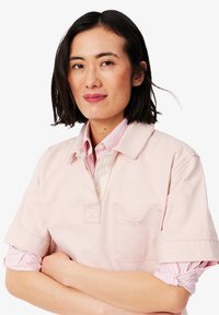 Rosa Poloshirt mit kurzen Ärmeln, Kragen und Tasche, versehen mit einem dezenten Logo. Darunter ist ein rot-weiß gestreiftes Hemd mit Kragen sichtbar.