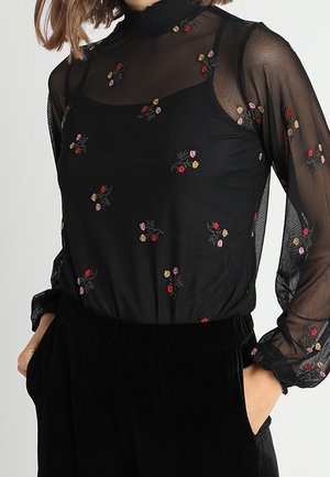 Sort gennemsigtig bluse med lange ærmer og høj hals med små broderede flerfarvede blomster, kombineret med sorte fløjlsbukser.