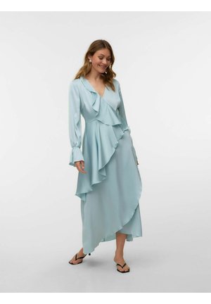 Frau lächelt, trägt ein hellblaues, langärmeliges Wickelkleid mit Rüschen und schwarze Sandaletten mit Absatz vor schlichtem Hintergrund.